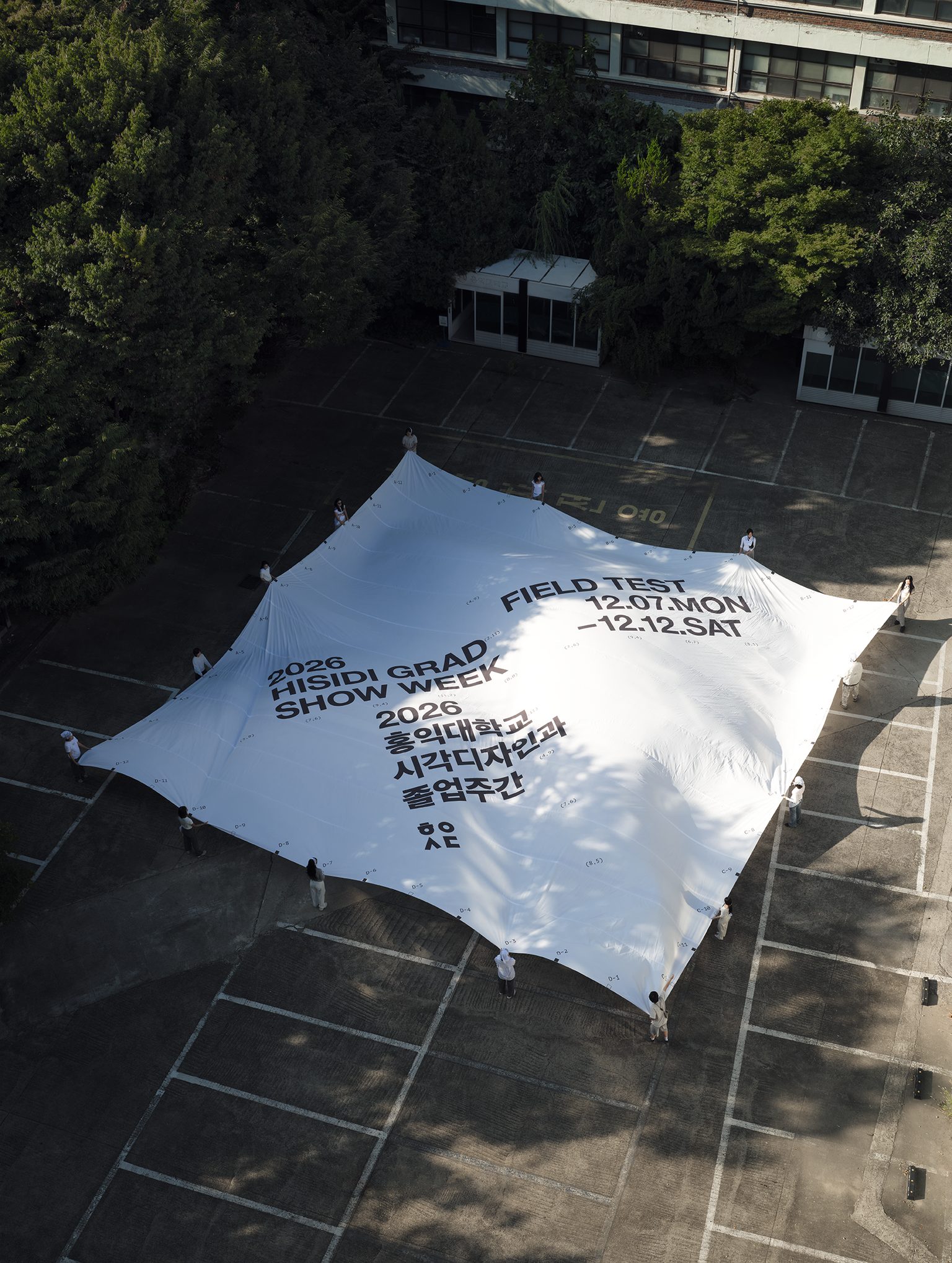 FIELD TEST 포스터 — 주차장 바닥에 펼친 커다란 흰 천을 사람들이 둘러 잡고 있다. 천 위에 2026 HISIDI GRAD SHOW WEEK, FIELD TEST 12.07.MON–12.12.SAT, 2026 홍익대학교 시각디자인과 졸업주간